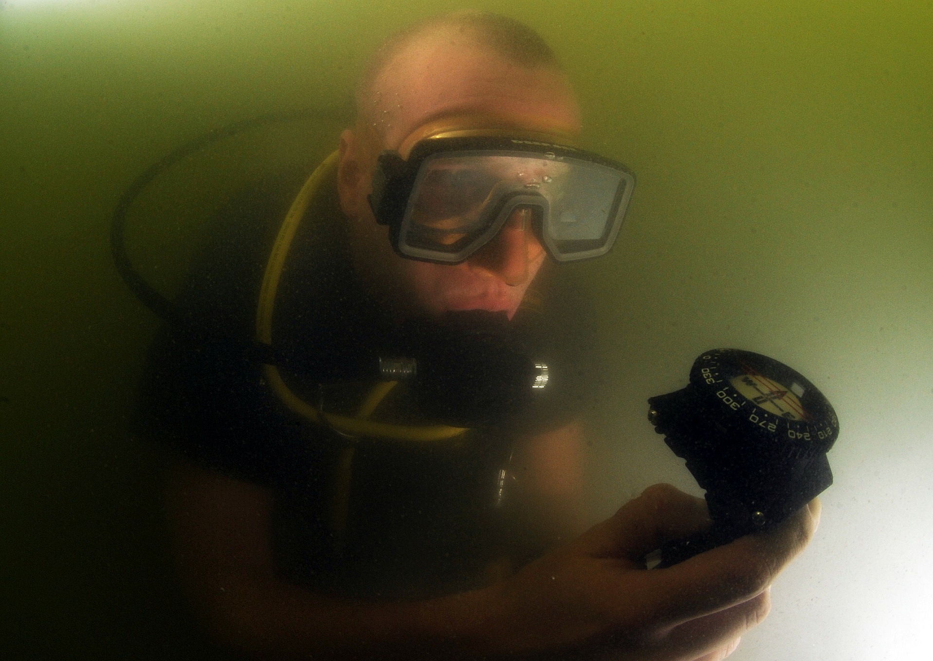 Taucher im Trübwasser hält einen Tauchkompass mit deutlich sichtbarer Gradskala vor sich und liest den Kurs ab.