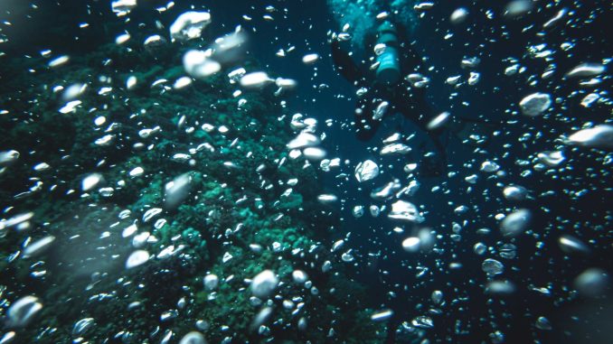 Dichte Wolke aus aufsteigenden Luftblasen im dunkelblauen Wasser, dahinter ein Taucher mit Flasche