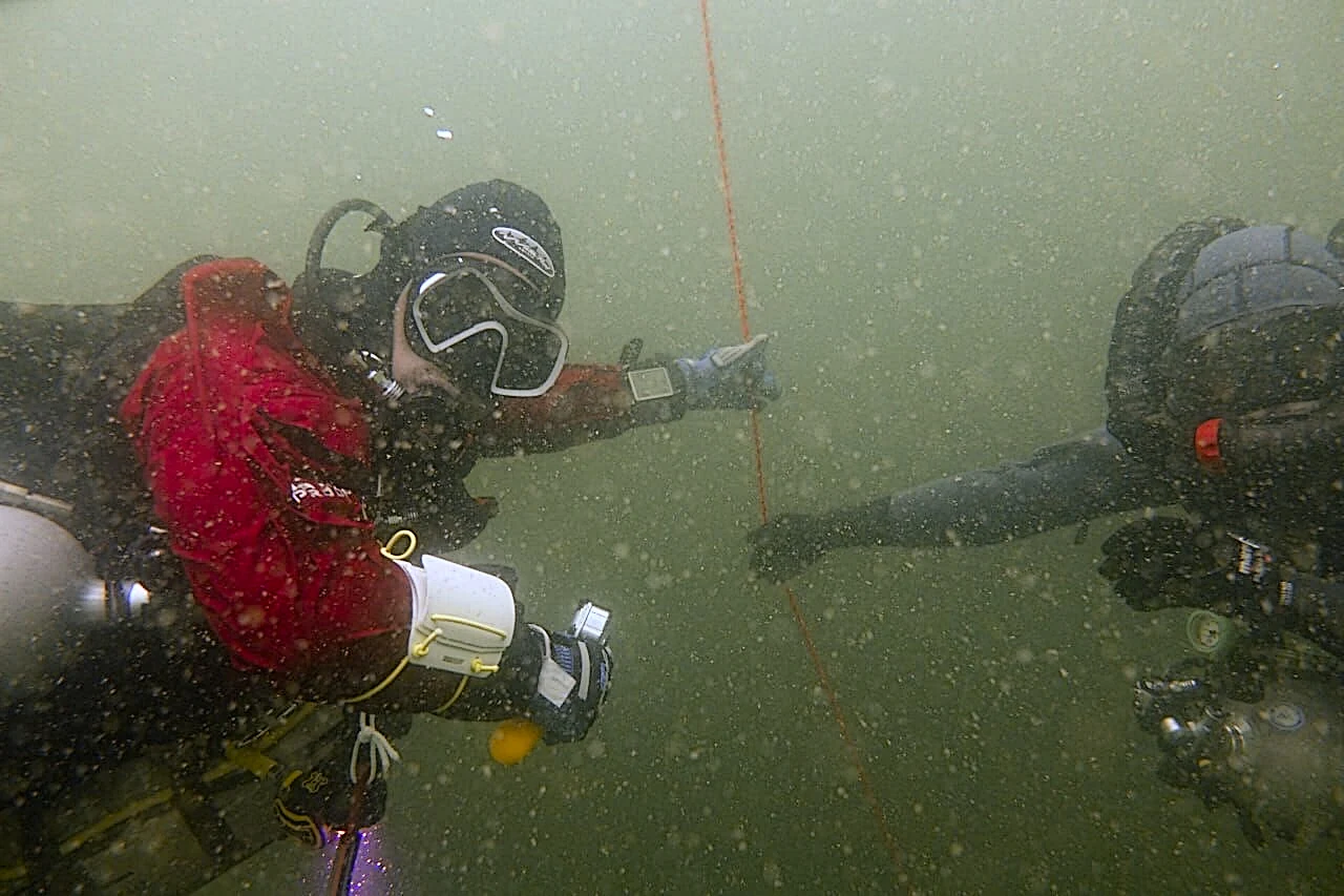 Zwei Taucher hängen im trüben Wasser nebeneinander an einer Leine und sitzen ihre Dekompressionsstufen ab, einer im roten Trockenanzug