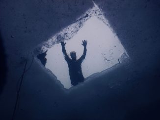 Blick von unten durch ein in die Eisdecke gesägtes Loch, darüber die Silhouette einer Person, links die Sicherungsleine