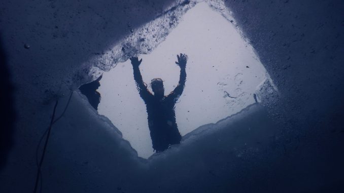 Blick von unten durch ein in die Eisdecke gesägtes Loch, darüber die Silhouette einer Person, links die Sicherungsleine