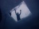 Blick von unten durch ein in die Eisdecke gesägtes Loch, darüber die Silhouette einer Person, links die Sicherungsleine
