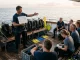 Tauchguide erklaert einer im Halbkreis sitzenden Gruppe an Bord den Tauchgang, mit Briefingtafel in der Hand