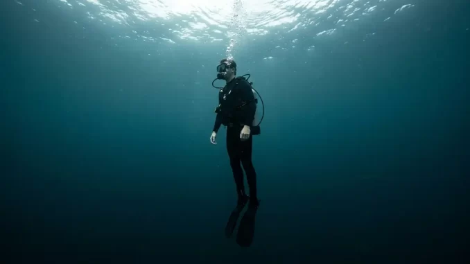 Vollständig ausgerüsteter Taucher sinkt mit entlüftetem Tarierjacket aufrecht und füße voran in tieferes Wasser ab.