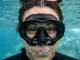 Gesicht einer Taucherin dicht unter der Wasseroberfläche hinter einer eng anliegenden Tauchmaske, darunter aufsteigende Luftblasen.