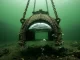 Eine unten offene Taucherglocke aus Metall hängt an Ketten unter Wasser, unter ihrer Decke steht ein silbrig spiegelndes Luftpolster.