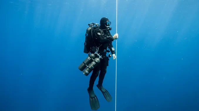 Technischer Taucher mit Doppelgeraet und zwei seitlich eingeklinkten Stageflaschen haengt an einer senkrechten Dekompressionsleine im Blauwasser.