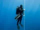 Technischer Taucher mit Doppelgeraet und zwei seitlich eingeklinkten Stageflaschen haengt an einer senkrechten Dekompressionsleine im Blauwasser.