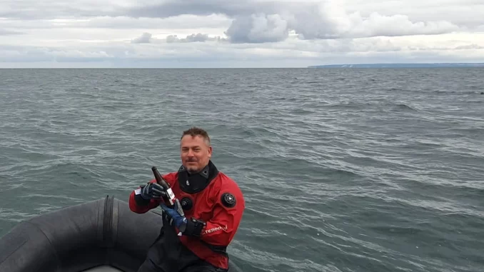 Taucher im roten Trockentauchanzug sitzt mit einer Bierflasche in der Hand im Schlauchboot auf der Ostsee, im Hintergrund die Küste von Rügen unter bedecktem Himmel