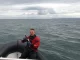 Taucher im roten Trockentauchanzug sitzt mit einer Bierflasche in der Hand im Schlauchboot auf der Ostsee, im Hintergrund die Küste von Rügen unter bedecktem Himmel