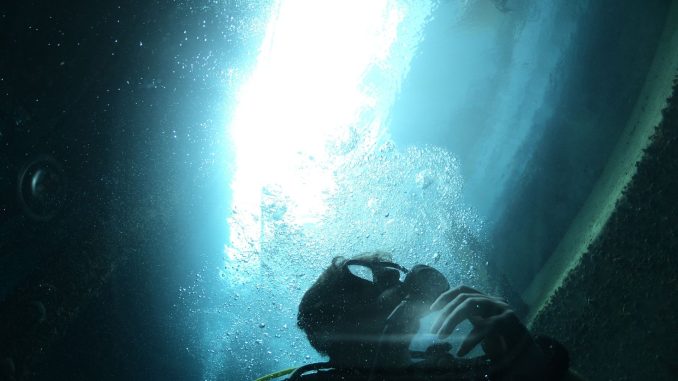 Taucher in dunklem Wasser vor einer gewölbten Wand, von oben fällt ein heller Lichtstreifen ein, Blasen steigen auf