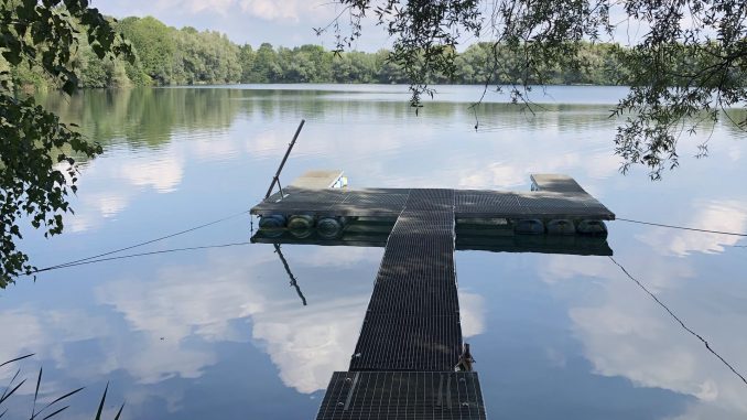 Gitterrost-Steg mit schwimmender Plattform auf einem ruhigen Baggersee, im Hintergrund bewaldetes Ufer
