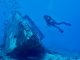 Taucher schwebt frei im blauen Wasser neben dem Bug eines auf Sand liegenden Schiffswracks, darüber ein Fischschwarm