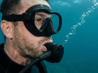Nahaufnahme eines Tauchers unter Wasser mit Atemregler im Mund, aus dem Ausatemventil steigt eine feine Blasenkette auf.
