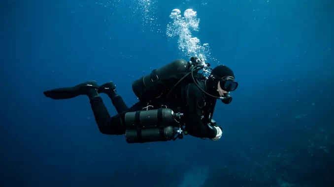 Technischer Taucher mit Doppelgeraet und zwei seitlich angeclippten Stageflaschen in tiefblauem Wasser.