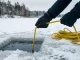 Behandschuhte Hände halten eine gelbe geflochtene Signalleine, die über die Kante eines Eisloches ins dunkle Wasser führt; daneben die aufgeschossene Leine