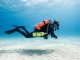 Einzelner Taucher schwebt waagerecht über hellem Sandgrund, seitlich an der Hüfte eine kleine Reserveflasche mit eigenem Atemregler; kein zweiter Taucher im Bild.