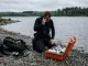 Taucher kniet am Seeufer neben einem geoeffneten Sauerstoffkoffer und telefoniert mit einem Mobiltelefon.