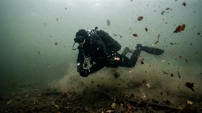 Taucher im Trockenanzug in gleichmäßig trübem, graugrünem Wasser eines durchmischten Herbstsees mit treibenden Laubblättern.