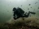 Taucher im Trockenanzug in gleichmäßig trübem, graugrünem Wasser eines durchmischten Herbstsees mit treibenden Laubblättern.